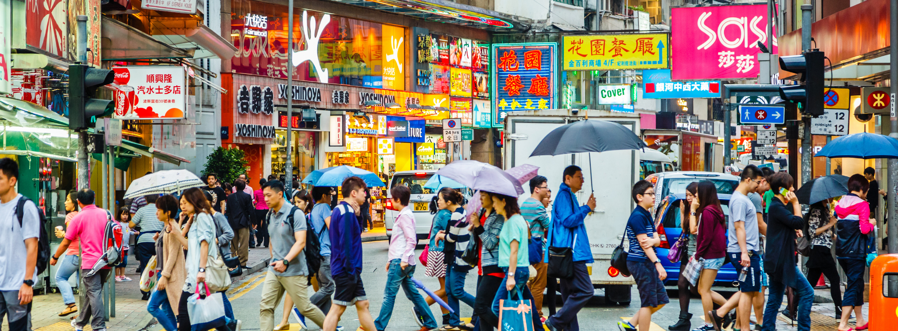 A busy and colourful street in Hong Kong with various people crossing a street, some holding umbrellas. 