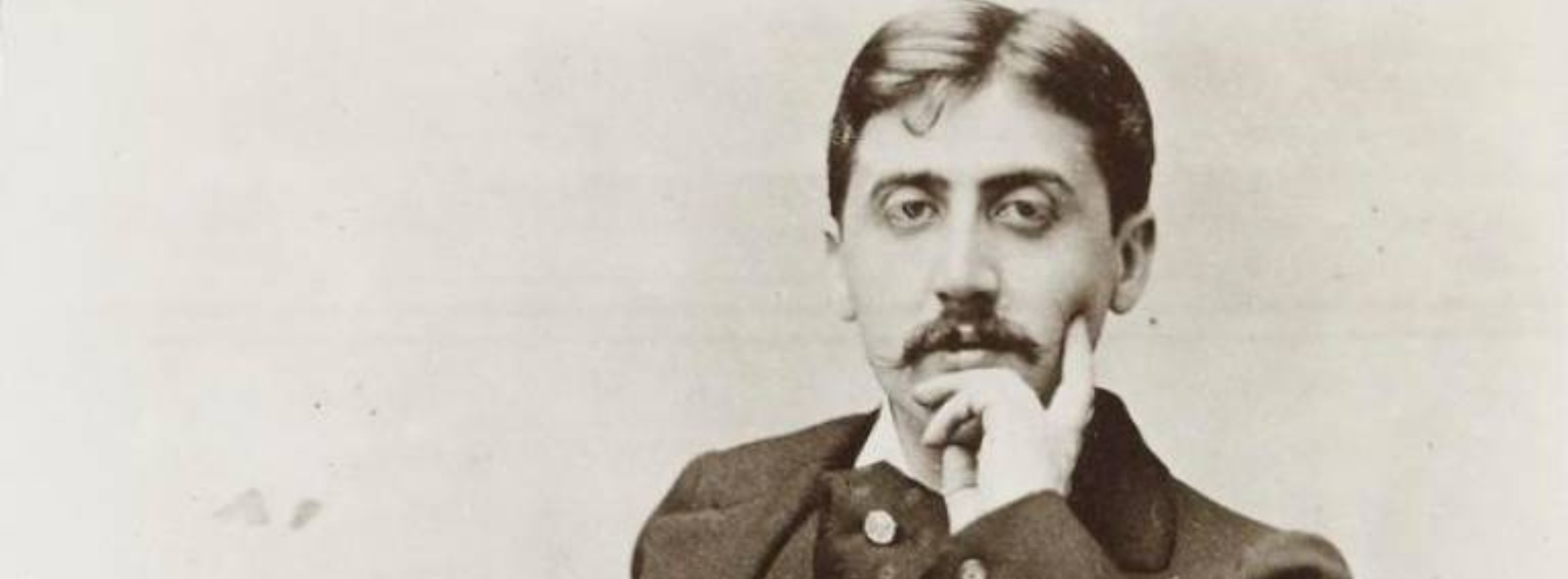 Marcel Proust looks to the camera, his chin in his hands. He is dressed in a dark suit. His hair is well-kept, slicked back with one small curl falling over his forehead. 