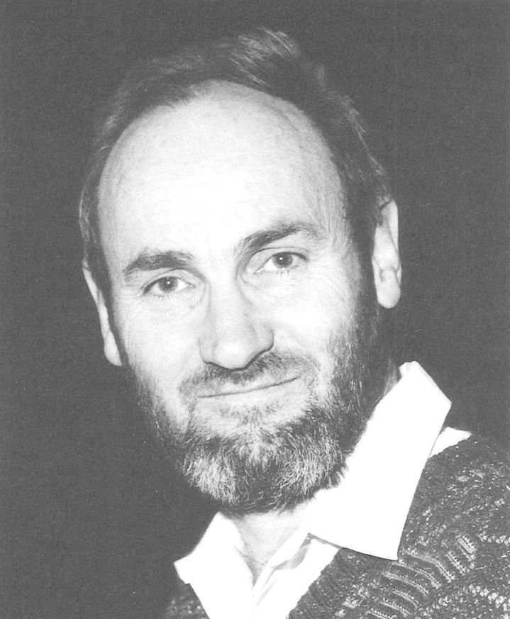 A portrait of John Oastler Ward, black and white. A man with a short beard wears a white collared top underneath a knitted jumper. 