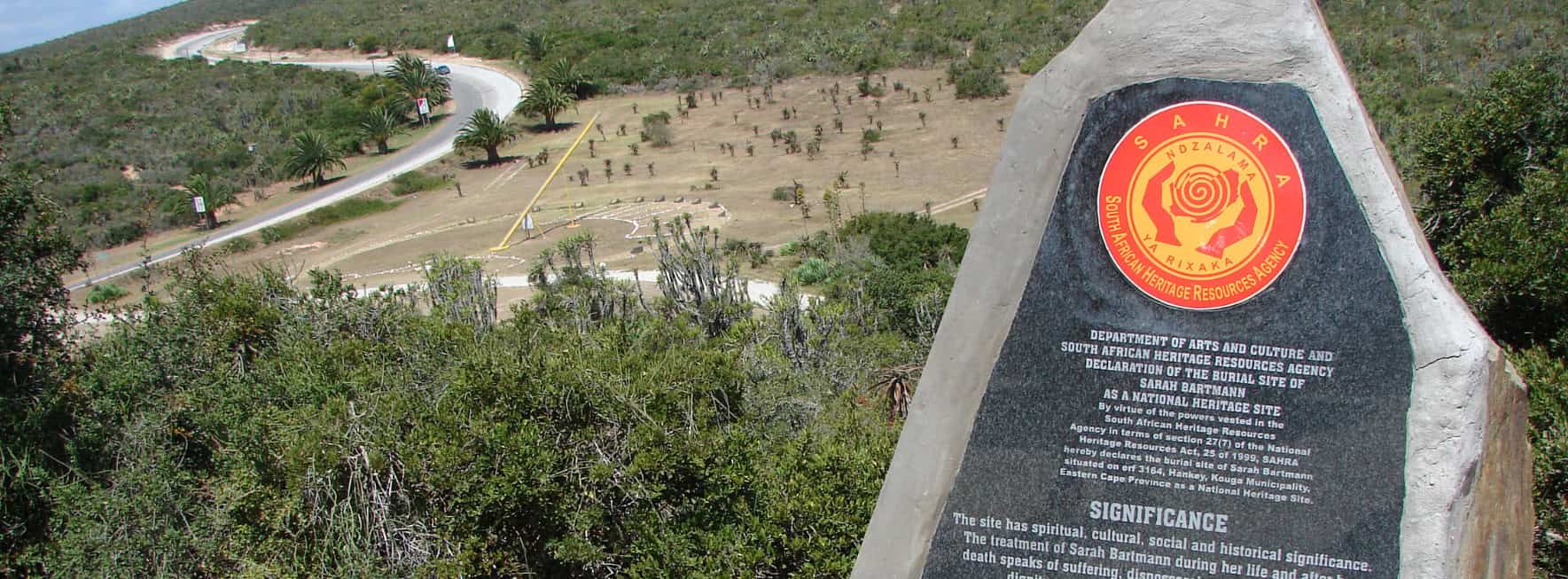 A grey memorial stone sits at the top of a hill. The plaque describes that the stone was erected in the memory of Sara Baartman.