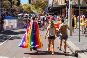 Three figures walk down a festive and bright Sydney street during Mardi Gras. Their backs are facing the camera. One wears a rainbow flag over their shoulders while another wears a rainbow hat. 