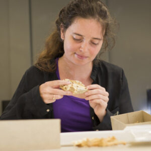 A woman with long brown hair carefully examines a small animal skull in a lab environment. She wears a black cardigan and a purple shirt. 
