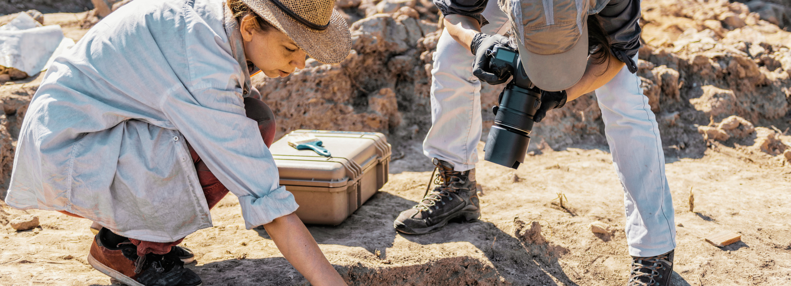 An image of a archaeologist working on a site. Beside her, a man photographs the excavation site. 