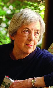 A distinguished woman in her eighties with short grey hair sits on a sofa chair. She wears a dark grey jumper, a delicate gold watch, and is looking out into the distance. 