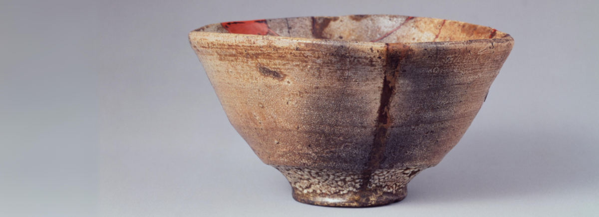 A light brown tea bowl from a museum. There is a long, dark brown line down the tea bowl indicating where it has cracked and been repaired.