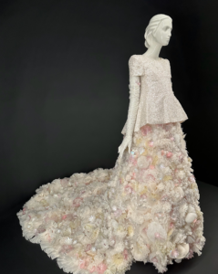 A mannequin at the Met Gala exhibiting a bridal-style skirt adorned with organza and tulle flowers in light pink and yellow flowers, with a peplum silk top. 