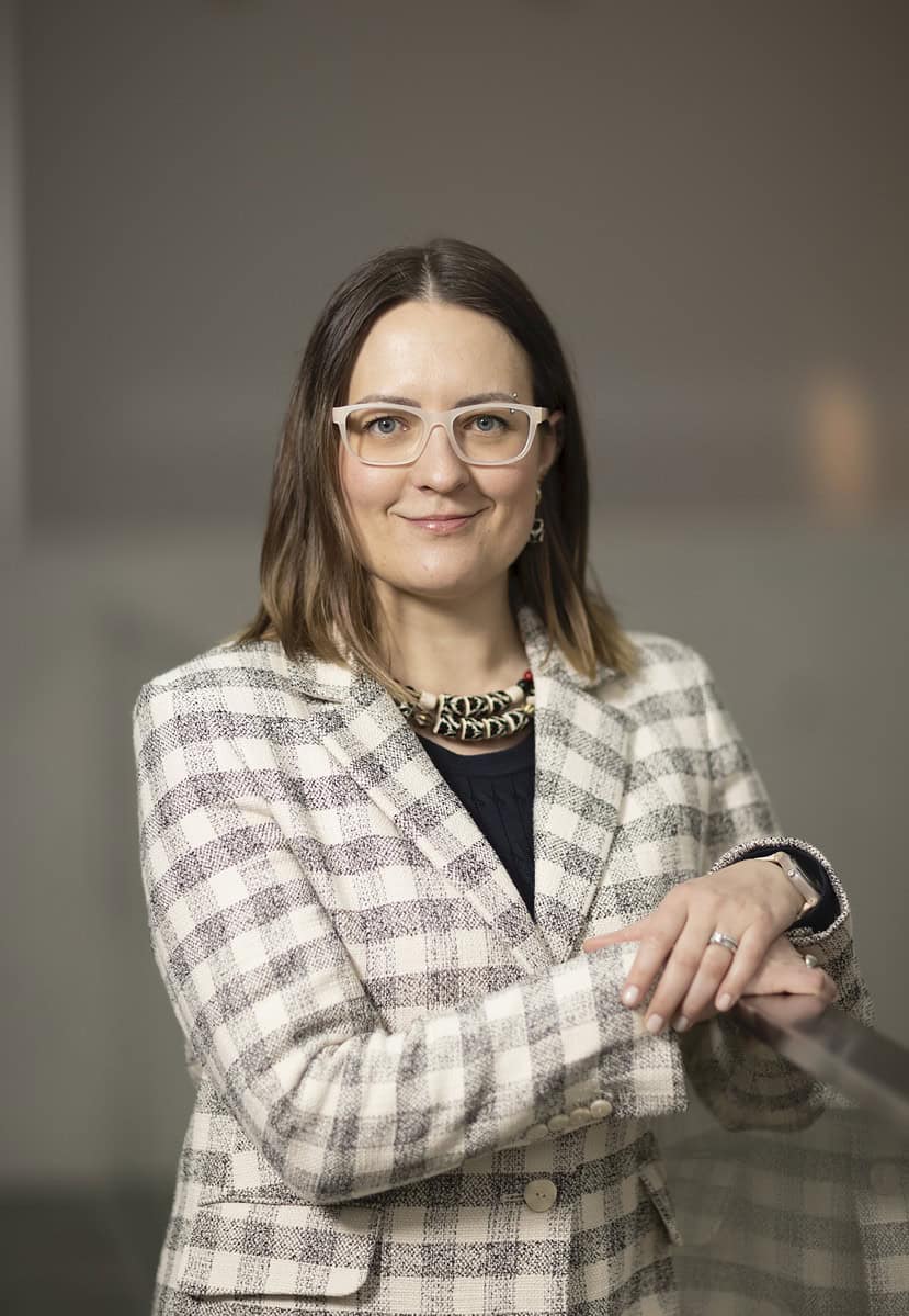 A young woman with brown hair poses for a photo. She wears a white and grey chequered jacket with tan glasses. 