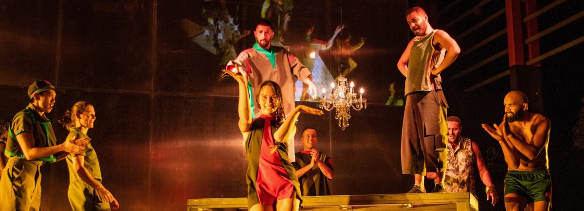 A photograph of a group of Marrugeku performers on a dimly lit stage. In the foreground, a woman is clapping, while other actors clap around her. 