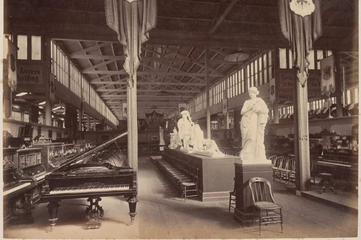 A black and white image of an international exhibition, showing a piano beside a marble statue inside a large hall, dated in the late 19th century.