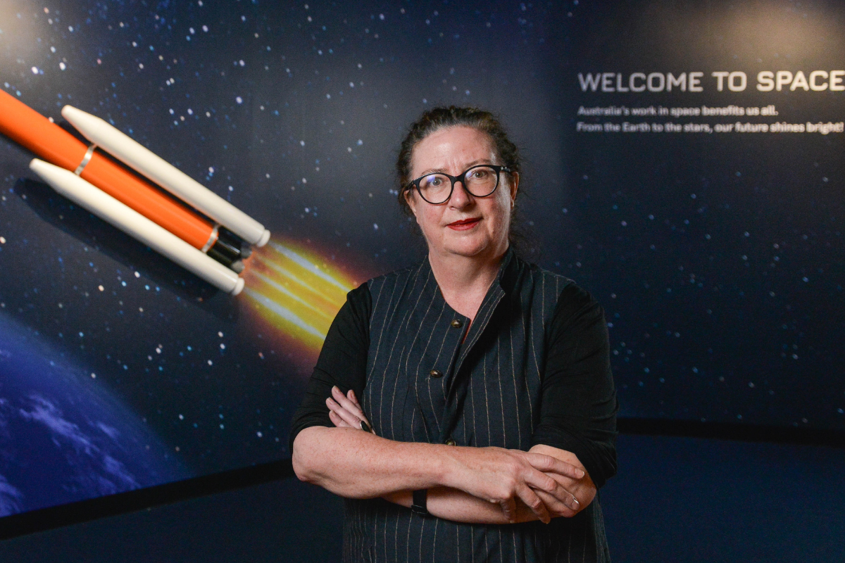 A woman stands with her arms crossed in front of a sign of a rocket with the words "welcome to space" printed beside the launching rocket.